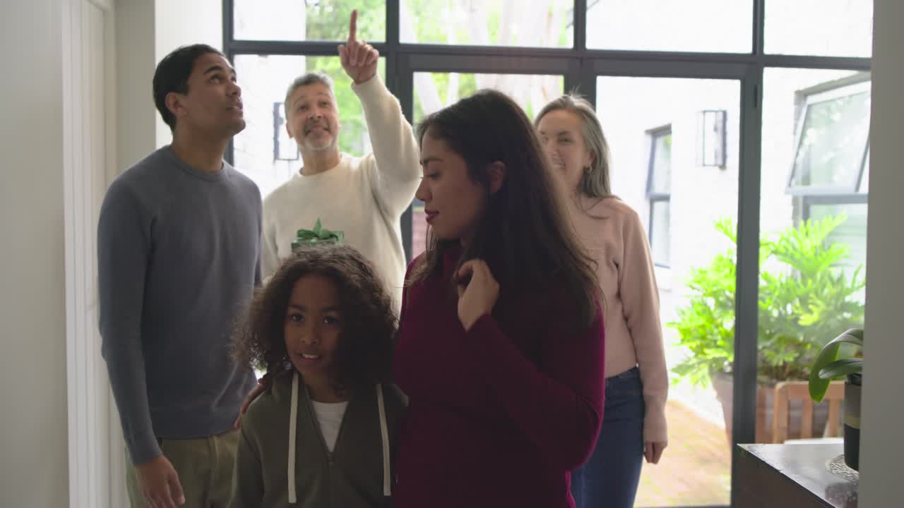 Family welcoming returning son arrival at foyer, offering green gift box and pointing ceiling light