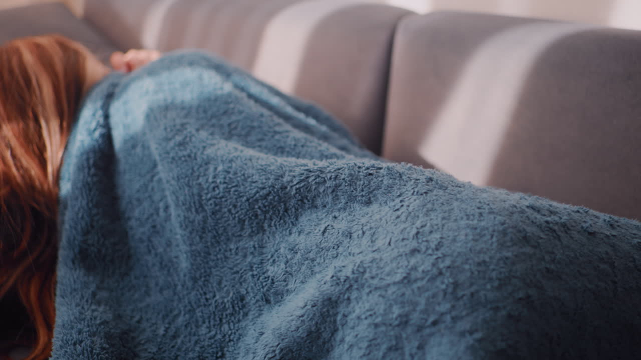 Close up of young woman facing couch while hiding under soft blue blanket, body fully tucked in warmth, sunlight falling across upholstery as she curls up quietly in serene resting position indoors