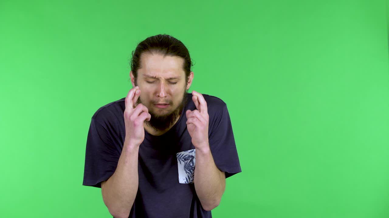 Caucasian hopeful, excited man holds his fingers crossed and makes a wish, upset when it does not come true. Bearded guy in a black Tshirt in the studio on a green screen. Close up