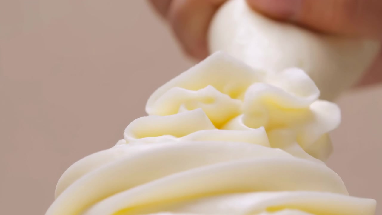 Primo piano che mostra una sac à poche tenuta in mano premuta per estrudere crema alla vaniglia chiara in rosette impilate e volute fluide, creando creste lucide, punte delicate e tenui riflessi gialli.