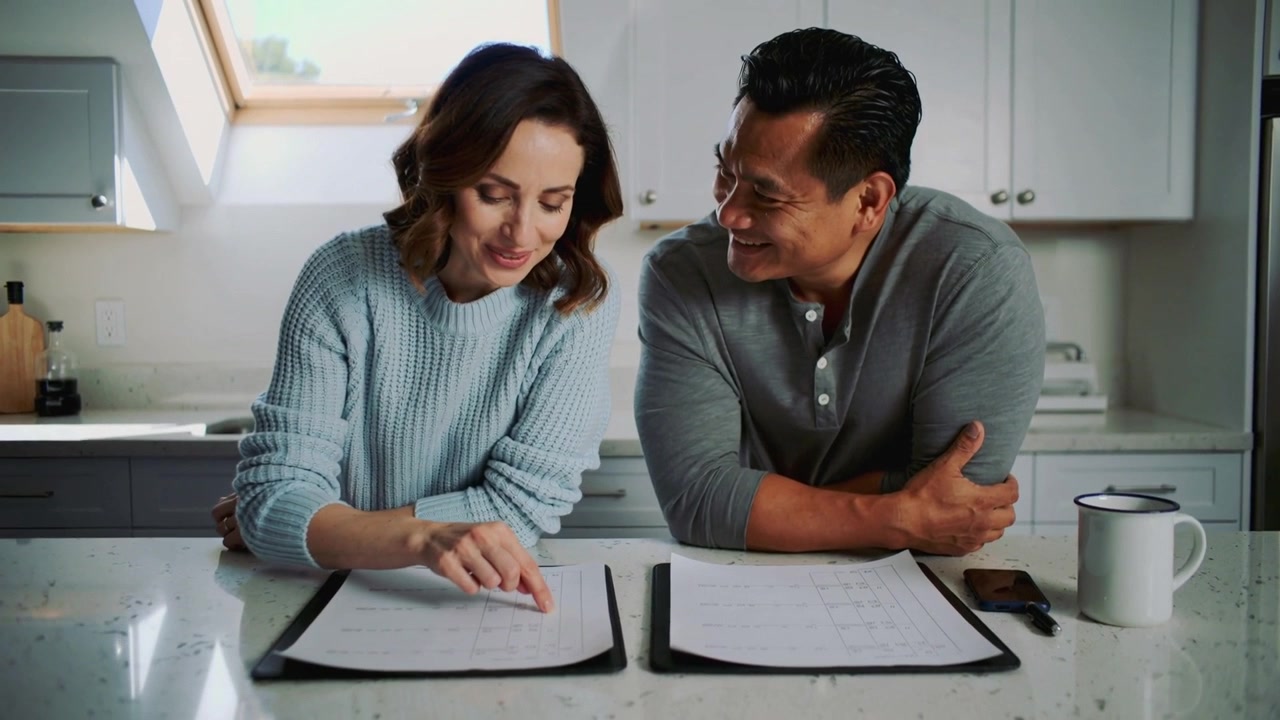 Donna che indica con un maglione blu, uomo sorridente con una maglietta henley grigia che esamina appunti al bancone della cucina