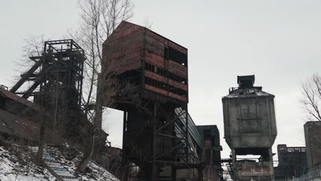 Zona-Industrial-Desolada-Y-Abandonada-Con-Grandes-Estructuras-Metálicas-Oxidadas,-árboles-Desnudos-Y-Nieve-En-El-Suelo,-Frente-A-Un-Cielo-Nublado