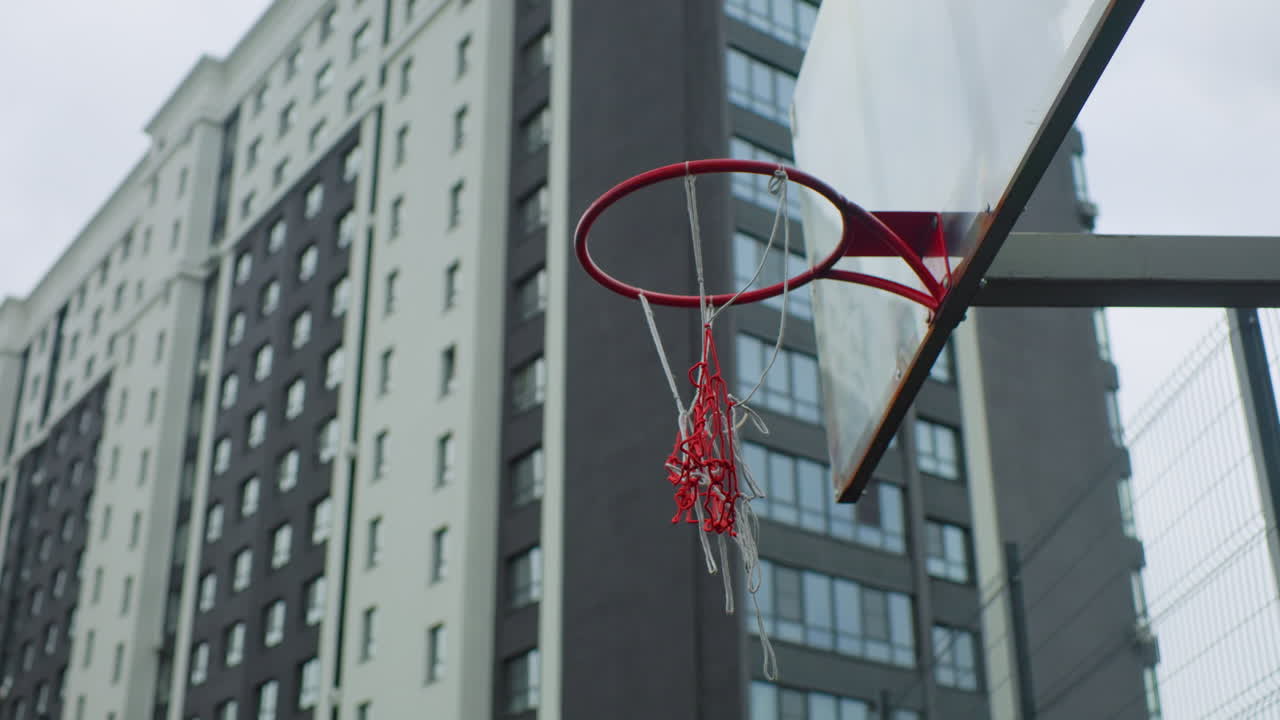 canestro da basket in città con grattacieli sullo sfondo, rete logora, tabellone angolato, recinzione in filo metallico, atmosfera da cortile grintoso, accenno di movimento, idea di allenamento, sfumatura di cultura giovanile, atmosfera piovosa