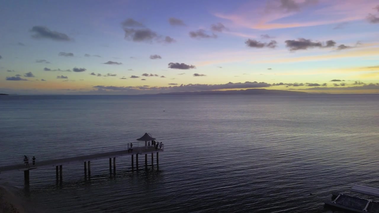 Betagende luftfoto af en træmole ved solnedgang med silhuetten af en ø i horisonten.
