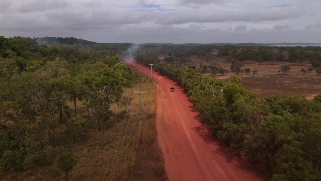 Luftfoto af et køretøj, der kører ad en støvet rød grusvej i den fjerntliggende australske outback, med en lille græsbrand, der brænder ved siden af vejen. Koncept for regional transport, risiko for skovbrand og fjerntliggende landskab.