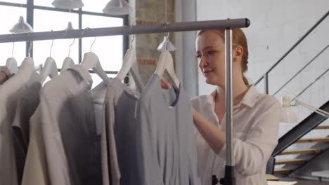 diseñadora de moda femenina mirando los vestidos en el perchero y luego mirando la cámara y sonriendo mientras trabajaba en el taller de costura