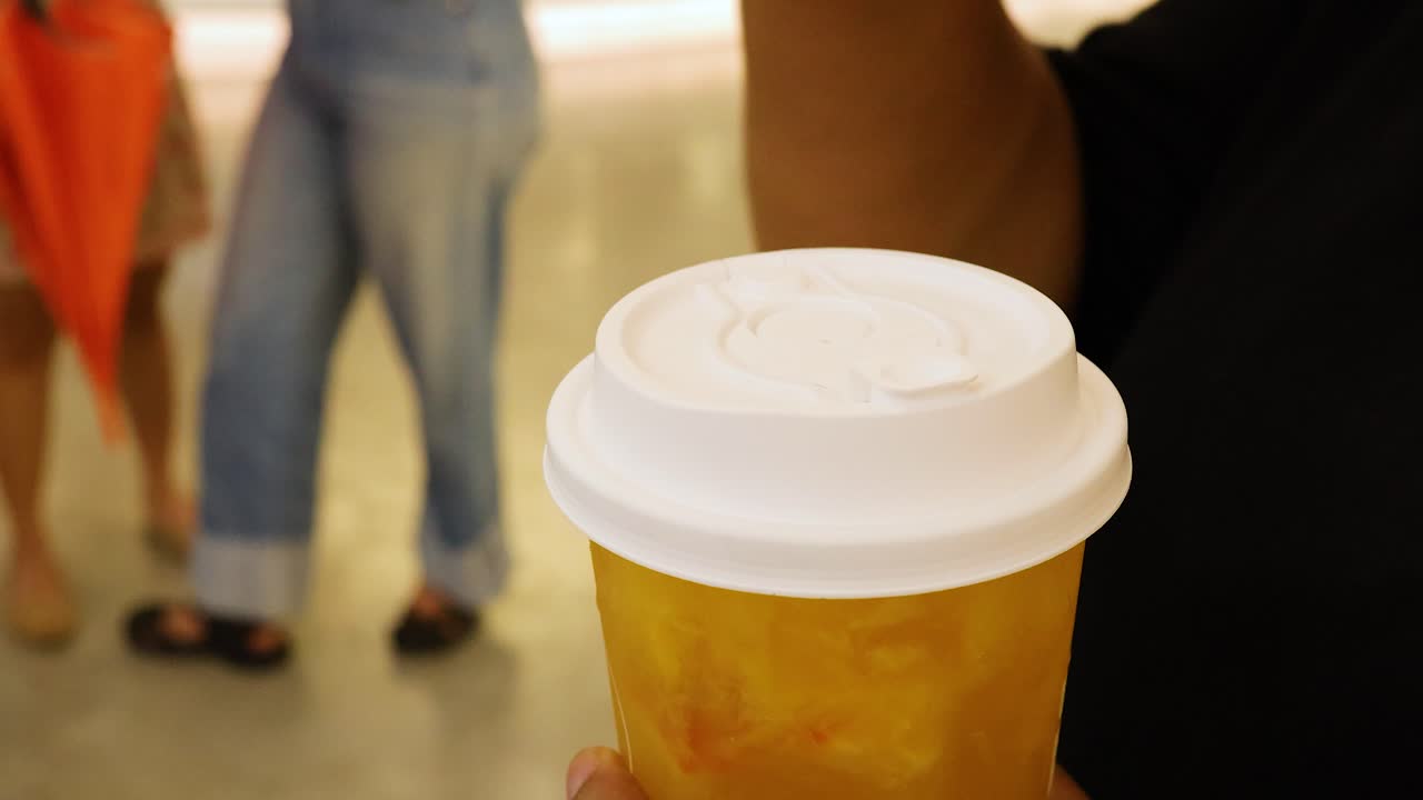 A hand opens a lid and inserts a straw into a fruit juice cup. Bright indoor setting with casual background