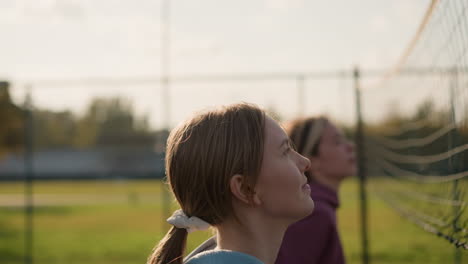 close up of youngster jumping during intense volleyball training session with net in front, with sunlight reflecting off hair, background features outdoor sports facility