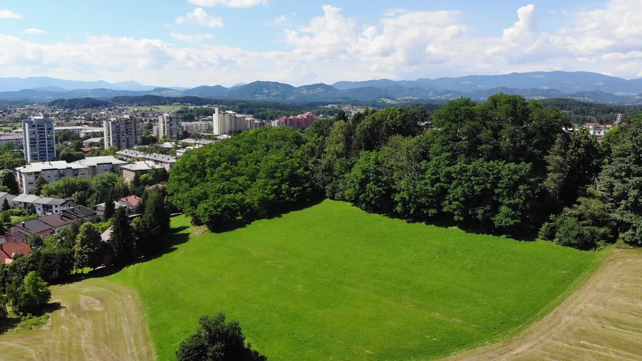 Aerial Over Grass Field Surrounded By Trees With Pan Left Reveal Of Town Of Celje In Slovenia