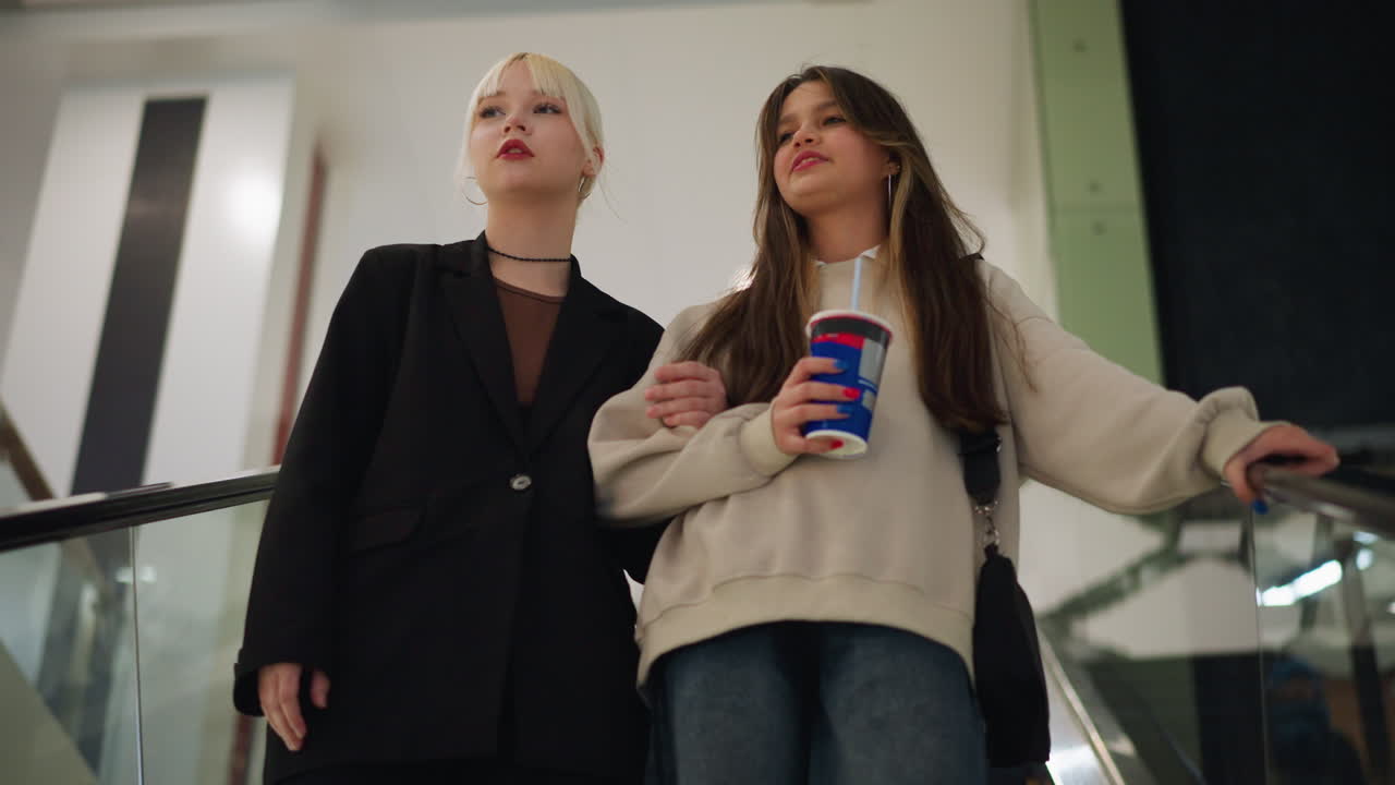 Upward view of ladies interacting and laughing while descending escalator inside shopping mall, holding drink cup, enjoying leisure time, sharing conversation and creating friendly joyful atmosphere
