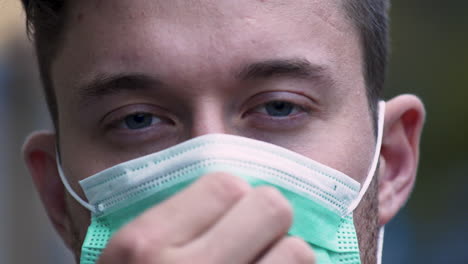 a young man with bright blue eyes and a stubble puts on a green protective covid-19 facemask in public, adjusting it on his face, while looking straight into the camera, static close up 4k shot