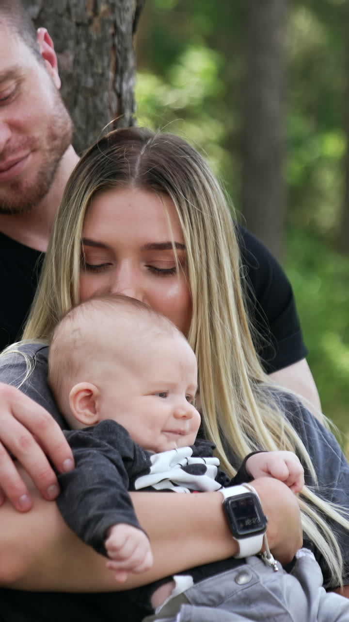 Kaukasische Familie zu dritt sitzt im Wald. Frau küsst ihr Baby auf den Kopf. Vertikales Video.