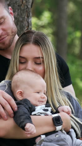 caucasian family of three sitting in the woods. woman kisses her baby to a head. vertical video
