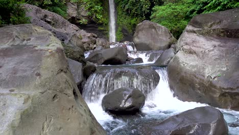 close up of river water rushing over boulders and rocks with a tropical waterfall in the background in a rainforest in asia