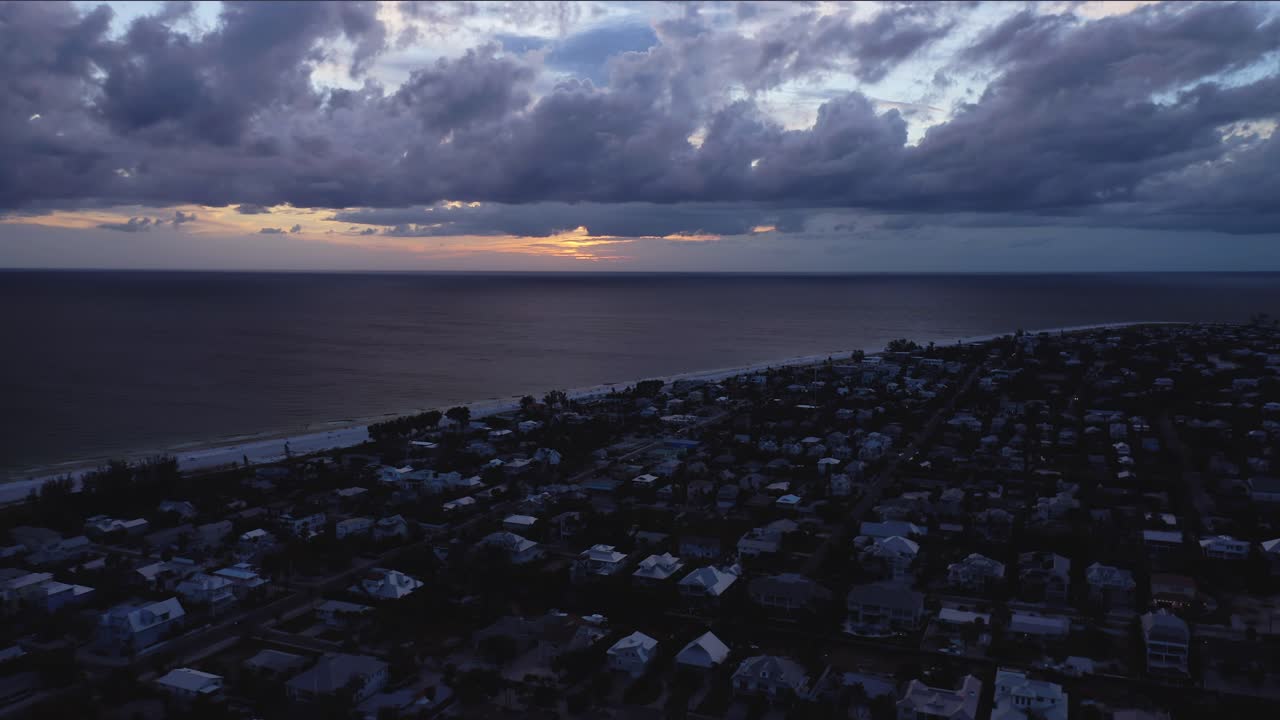 Dark rooftops line a quiet seaside town as a bruised sky thickens, a thin band of sunset glows over calm water, hinting at oncoming weather and restful hush