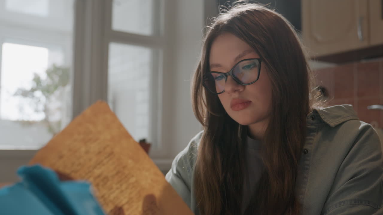 Jovem mulher lendo livro na cozinha, luz da janela e óculos, expressão focada revisando anotações e documentos, suéter casual e planta em vaso no parapeito da janela, ambiente matinal calmo com calor cinematográfico.