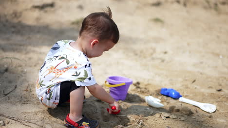 smiling baby boy squatted to play in sand. kid uses toy shovel to pick some sand.