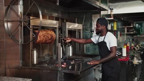 Un-Hombre-Con-Un-Delantal-Negro-Está-Haciendo-Girar-Una-Brocheta-De-Kebab-En-Un-Mercado-De-Doners.-Cocinar-Carne-Según-Los-Estándares-Orientales.-Vídeo-Filmado-En-Alta-Calidad