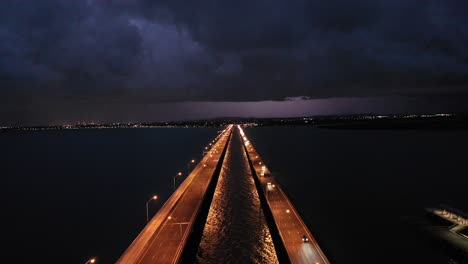 stationary drone shot of bridge and ocean with storm in the distance, lightning striking the town in distance, taken at night