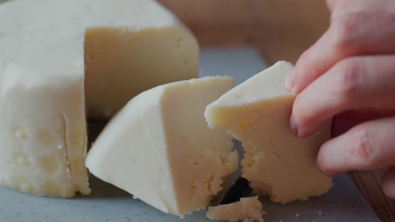 Una mano corta cuidadosamente una cuña de queso blando. El cuchillo se desliza suavemente a través de la textura cremosa. El queso tiene una superficie lisa y blanca. Los dedos de la persona sujetan la rebanada firmemente