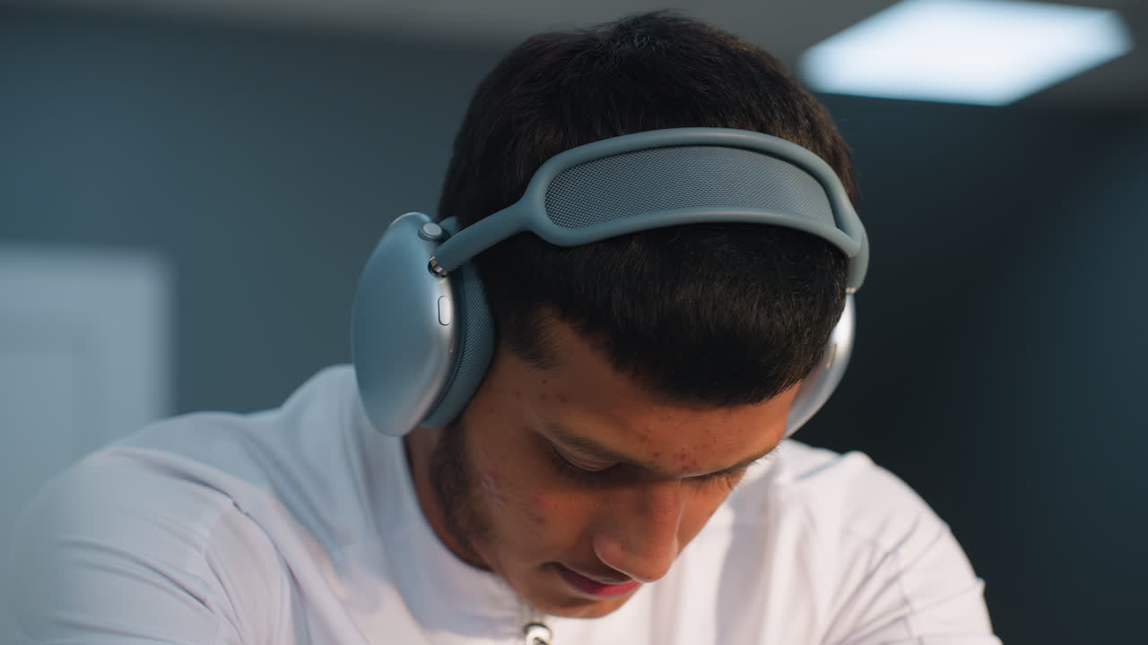 Close up of young man in white top wearing headphones, tilting head thoughtfully with soft expression, blurred gym interior and equipment in background under bright lights