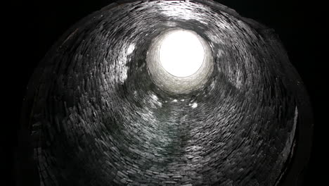 looking up from the bottom of a cylindrical brick shaft tunnel, with a bright circular opening at the top providing light