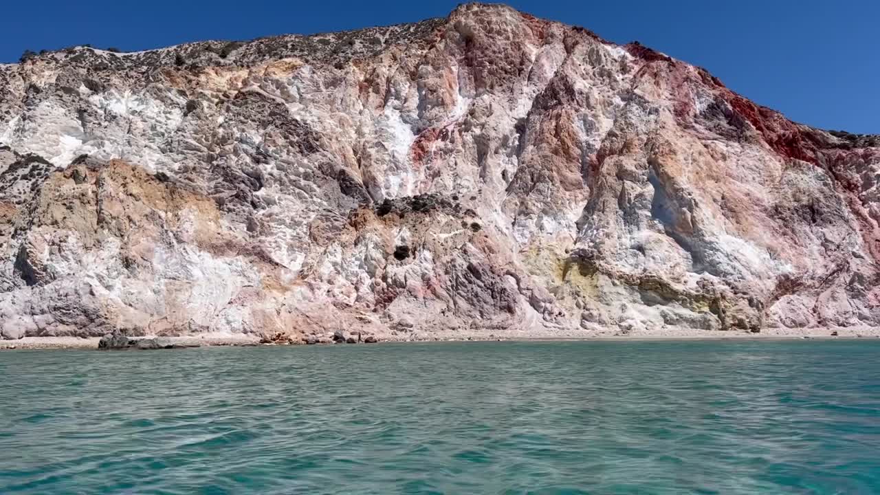 Vibrant cliffs in white, pink, yellow, and red rise above calm turquoise waters of the Aegean Sea under a bright blue sky on Milos Island, Greece.