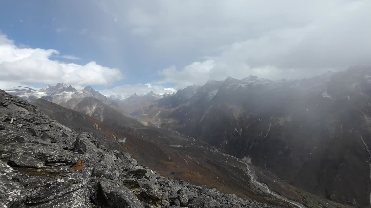 un panorama impresionante de un valle del himalaya nepalés se despliega mientras un punto de vista alto captura la dramática interacción de picos cubertos de nieve, nubes giratorias y una suave nevada.
