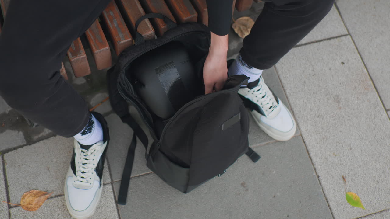 sitting on bench opening backpack, hands check interior pockets and straps while white sneakers rest on pavement, quiet autumn leaves nearby, casual outfit with hoodie and jacket, minimalist city