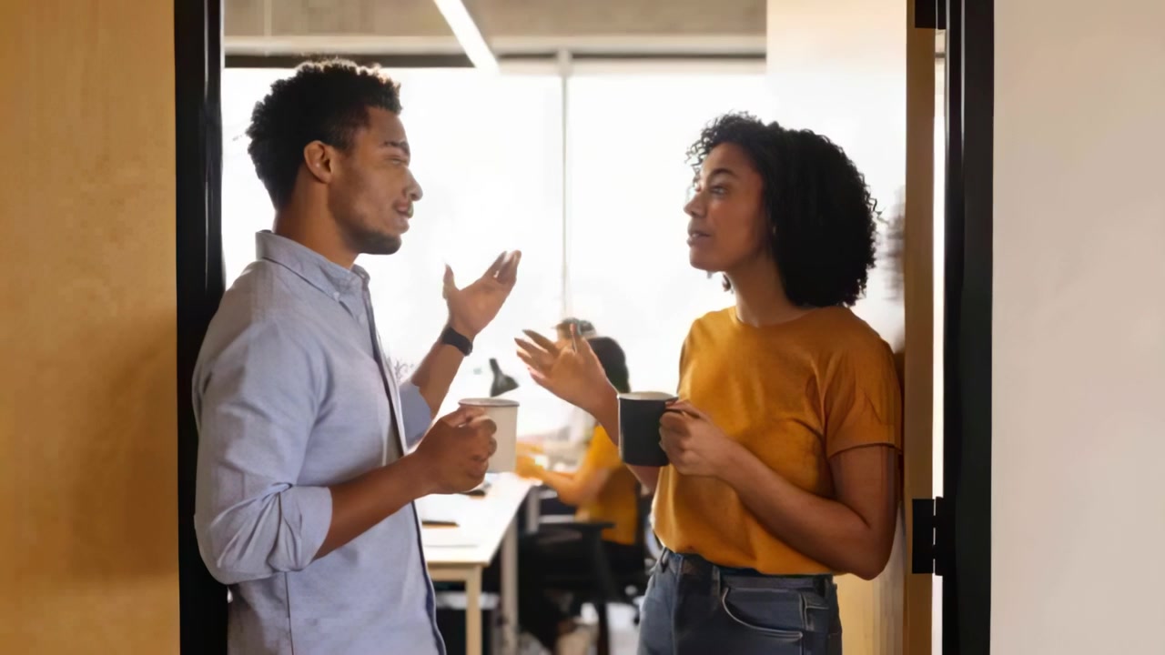 Meeting at office doorway, two exchanging notes, holding mugs, woman in yellow top checking phone