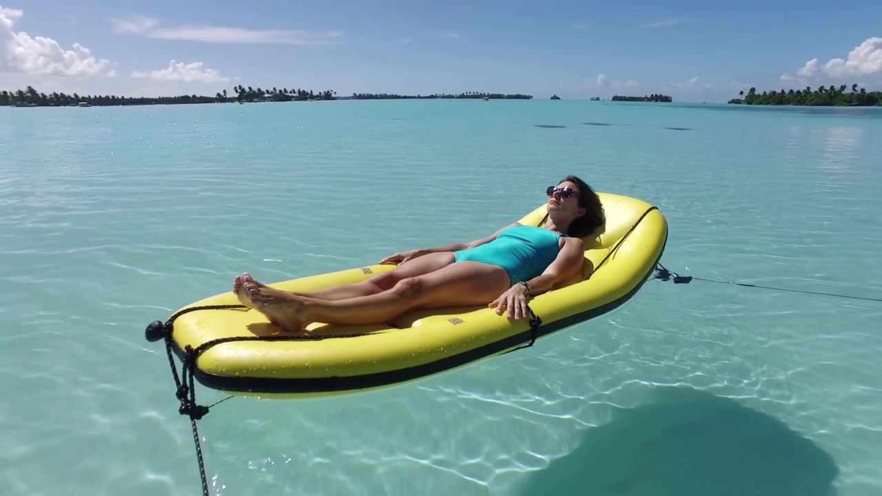 Sunbathing woman in teal swimsuit and shades on yellow float tethered in bay, gentle breeze nudging