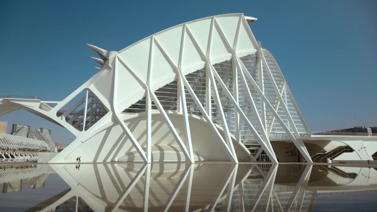 Edificio futuristico bianco di riferimento con guscio curvo e ampio, nervature di supporto diagonali, facciata in vetro e riflesso a specchio in acqua immobile sotto un cielo azzurro limpido, visto da un'ampia angolazione bassa e luminosa.