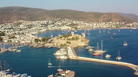 Luftdrohne-Der-Burg-Von-Bodrum,-Während-Ein-Boot-An-Einem-Sonnigen-Sommernachmittag-In-Der-Mugla-Türkei-In-Den-Jachthafen-In-Der-Mugla-Türkei-Einfährt,-Während-Die-Sonne-über-Den-Hügeln-Untergeht