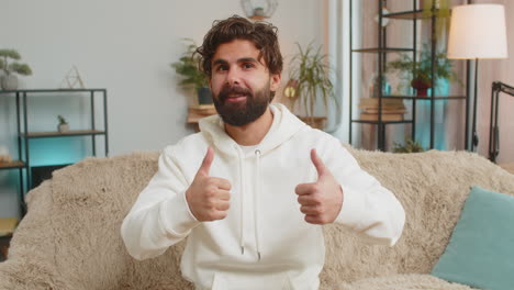 happy excited indian young man sitting on sofa couch looking at camera and showing thumbs up sign