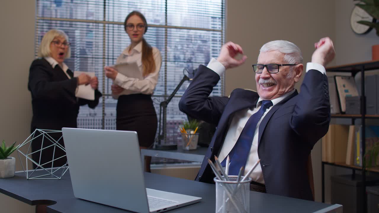 Cheerful mature old businessman raising hands celebrating sudden victory with colleagues in office