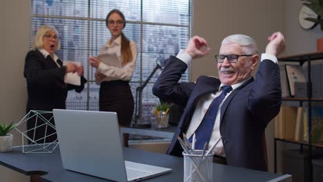 cheerful mature old businessman raising hands celebrating sudden victory with colleagues in office