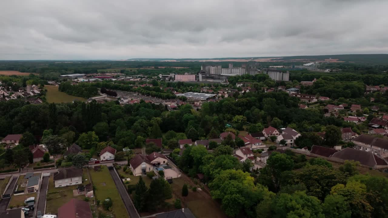 Slow aerial drone backward over quiet suburban village in the French countryside. Shows residential houses with gardens, trees, and apartment buildings in the distance under a cloudy sky