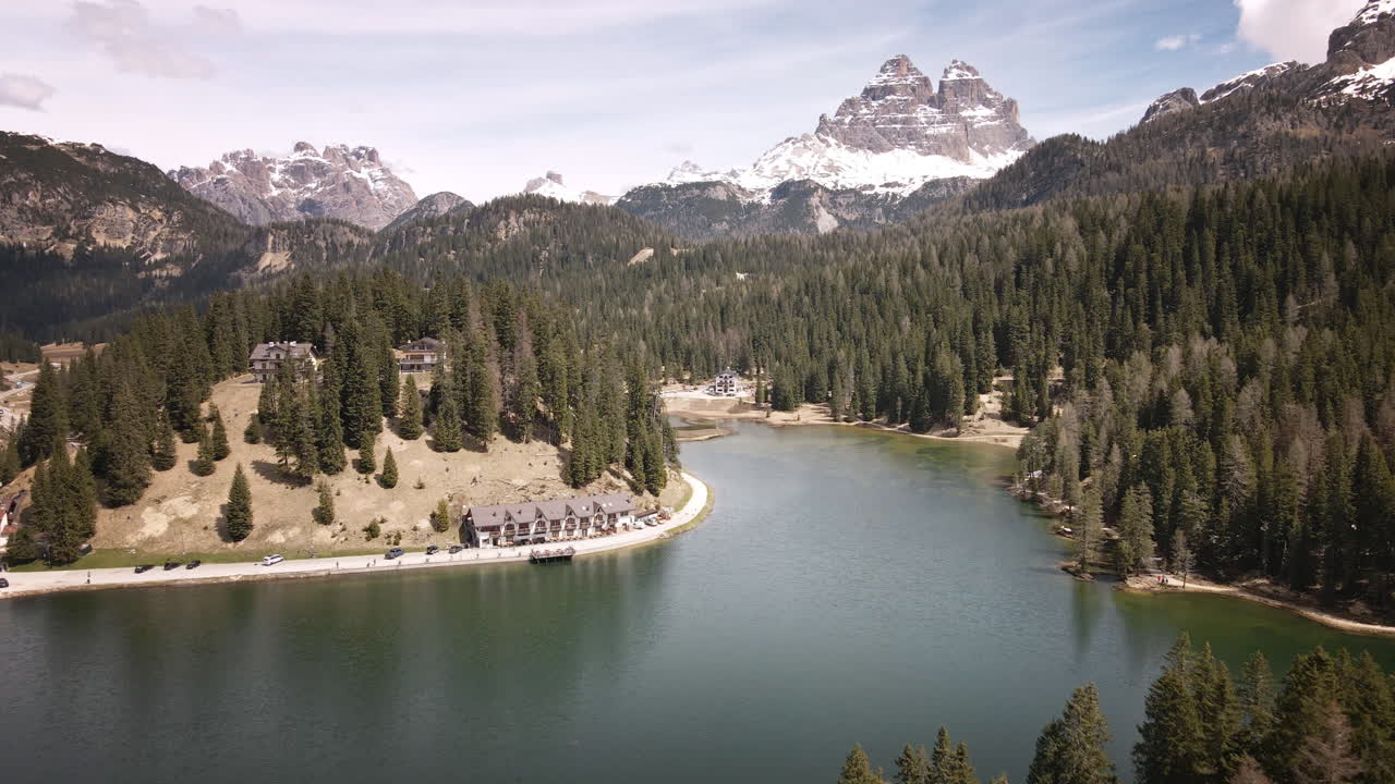 Camera moves forward above a calm alpine lake, passing evergreen forests toward snow‑capped Dolomite peaks and lakeside lodges on a bright spring day