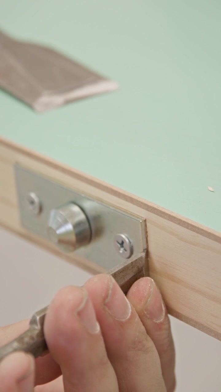 Vertical close-up of a carpenter chiseling a door edge to fit a metal hardware plate. Precision woodworking process refining a mortise for flush installation on a timber door.
