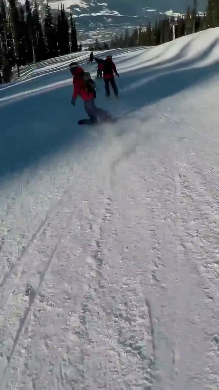 Vertical video: Carving boarder in red jacket going down run while camera following toward treeline