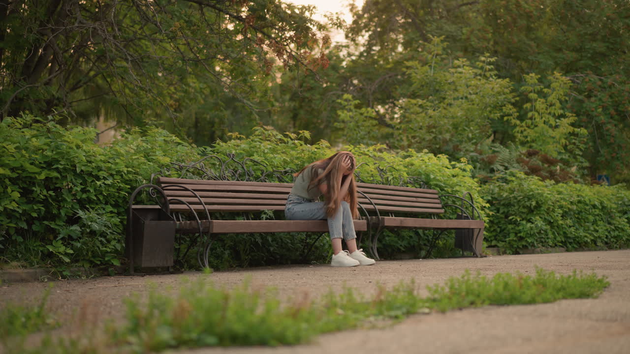 Lady seated on wooden bench in reflective, somber posture, hands gently holding her head and covering her face, long blonde hair cascades down her shoulder, surrounded by lush greenery