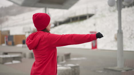 back view of woman stretching arms outdoors in winter setting with blurred people and light poles in background, showcasing serene workout routine in urban environment surrounded by light snowfall