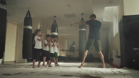 el entrenador muestra golpes a los niños en la escuela de deportes. los niños pequeños aprenden a golpear y dar pasos con un instructor hábil en el gimnasio a cámara lenta. combate de boxeo