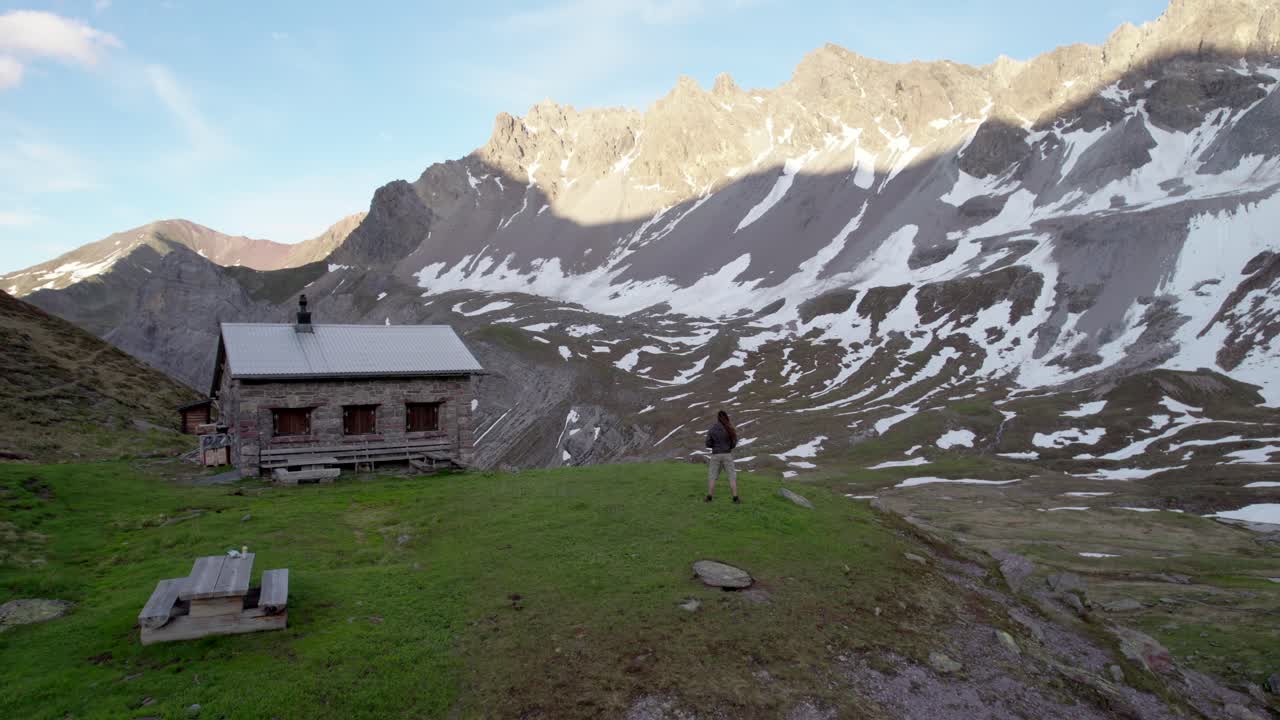 imágenes aéreas de drones sobrevolando a un hombre parado junto a una cabaña alpina suiza en un espectacular paisaje montañoso con parches residuales de nieve y prados alpinos en suiza