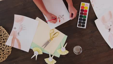 Cinematic-shot-of-a-girl-drawing-easter-bunny-in-a-white-paper-with-water-color-in-a-table