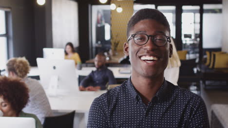 millennial black male creative wearing glasses smiling to camera in a busy casual office, close up