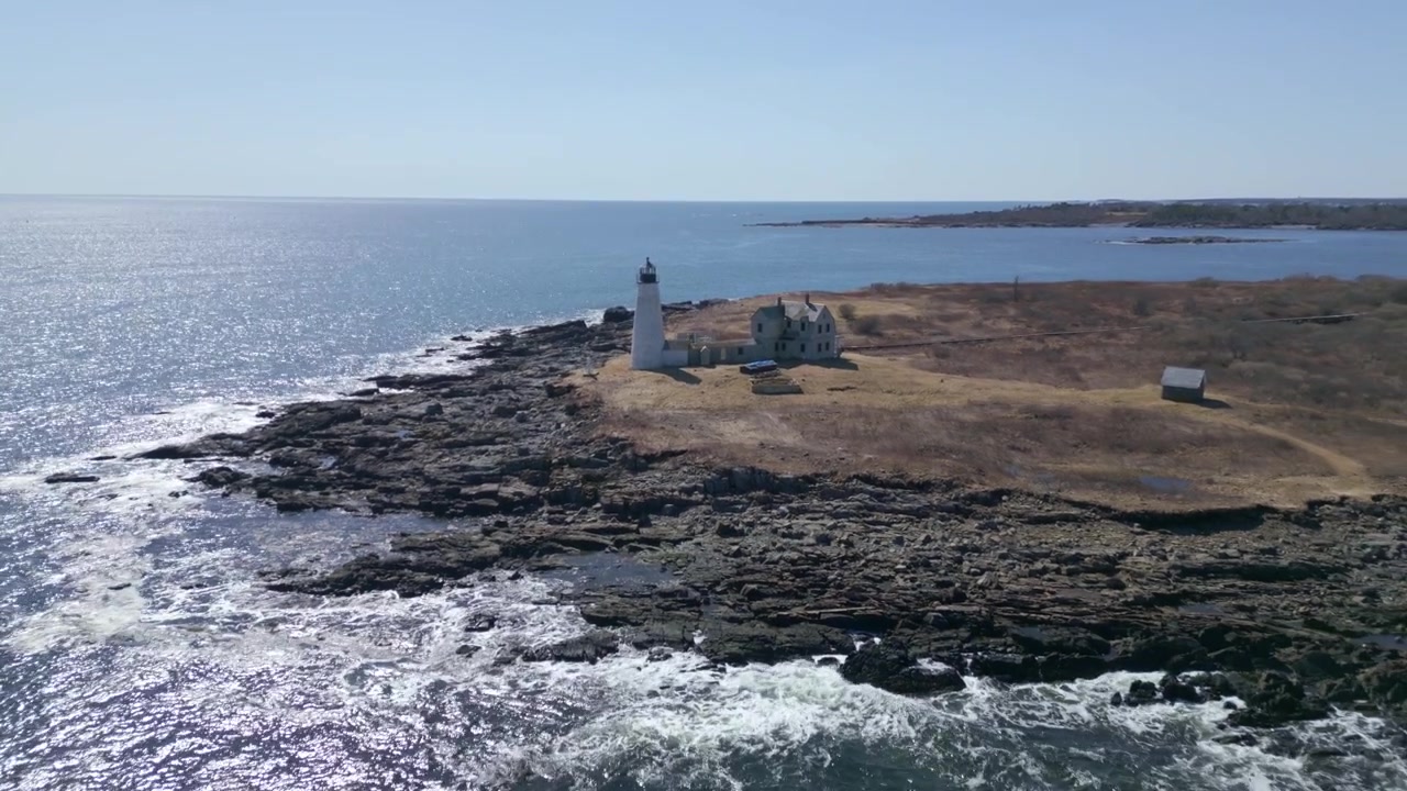 Luftfoto som trekker seg tilbake fra den robuste kystlinjen i New Hampshire med steinete strender og bølger som slår under et fyr