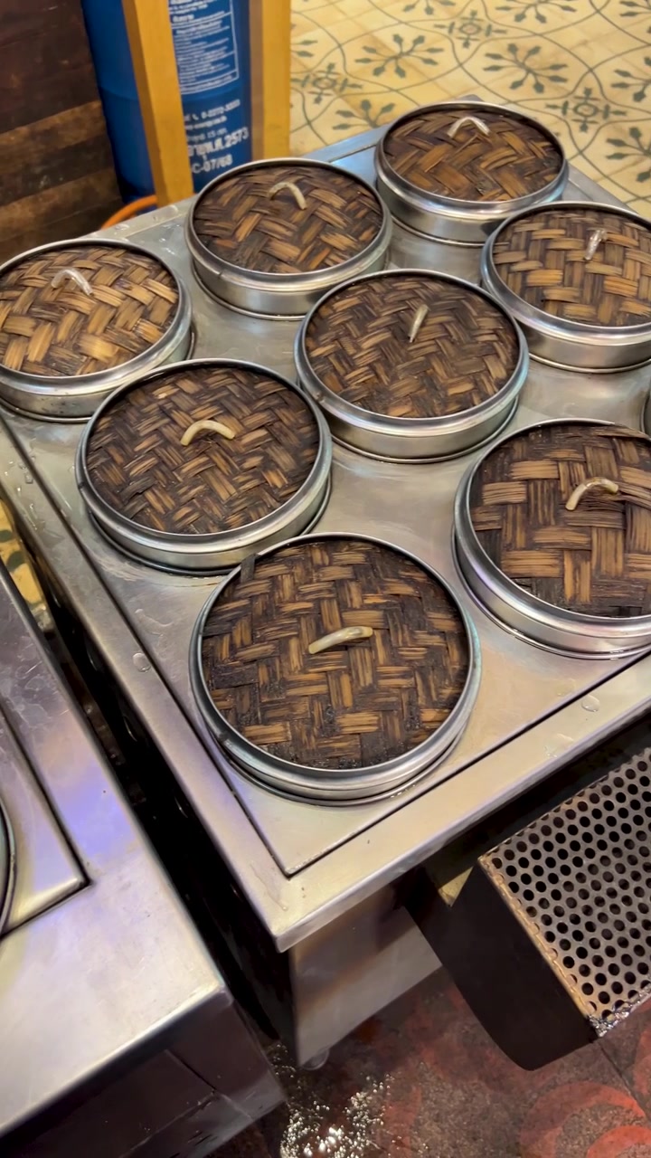 Camera circles stacked bamboo steamers on metal counter under warm lights at Bangkok street stall.