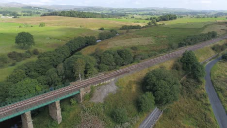 Drohnenaufnahmen,-Die-Sich-Parallel-Zu-Einer-Ländlichen-Eisenbahnlinie-über-Eine-Steinbrücke-Und-Einen-Fluss-Bewegen,-Umgeben-Von-Feldern,-Ackerland,-Hügeln-Und-Landschaft