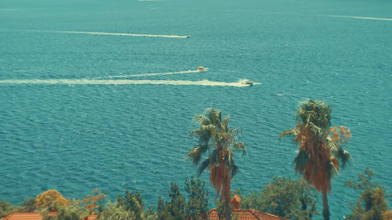 Mavi deniz suyuna bakan, beyaz izler bırakan teknelerin olduğu, ön planda bulanık çiçekler ve yeşil ağaçlarla çerçevelenmiş sinematik vintage görünüm